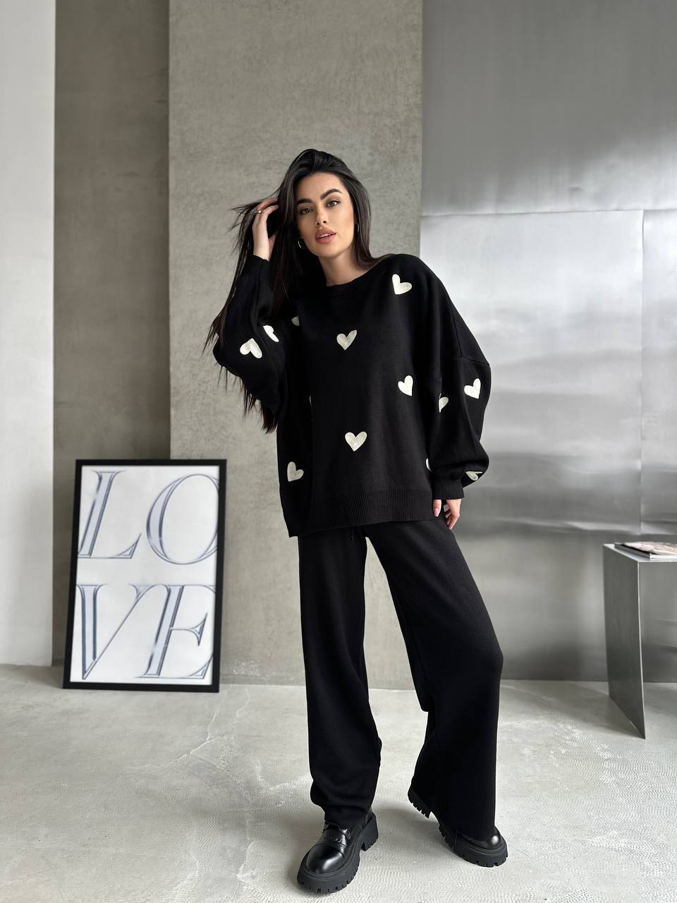Woman wearing a black outfit with heart patterns in a minimalistic room.
