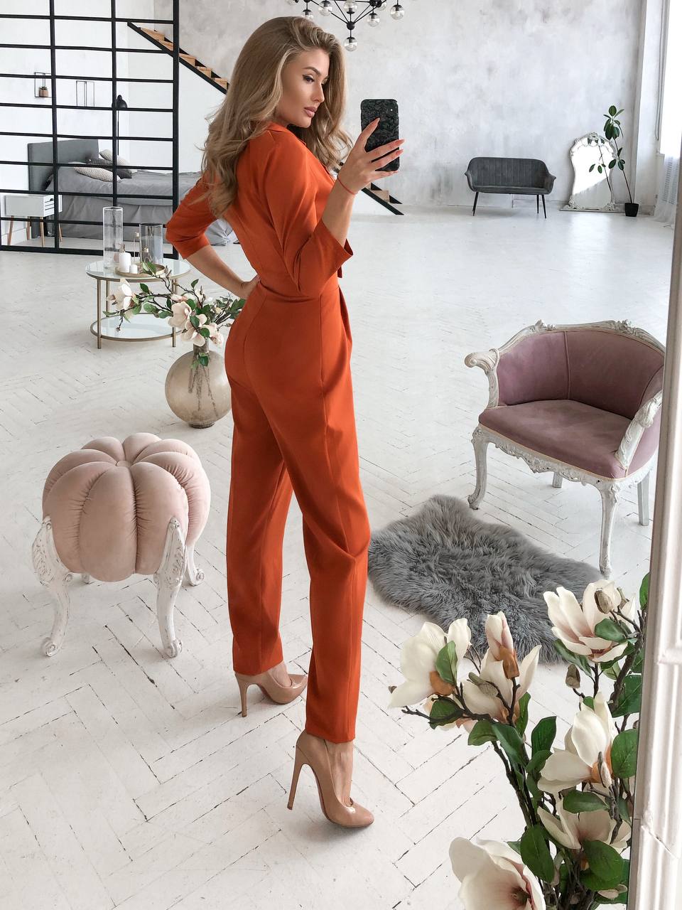 Woman in an orange jumpsuit taking a mirror selfie in a stylish room with decor elements.