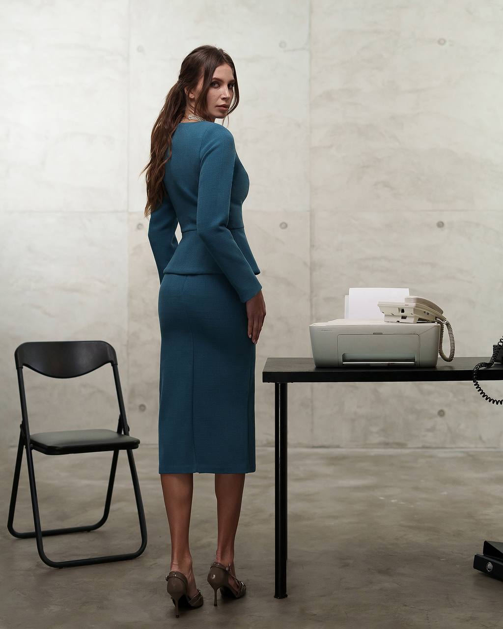 Woman in a teal dress standing in an office setting with a chair and printer.