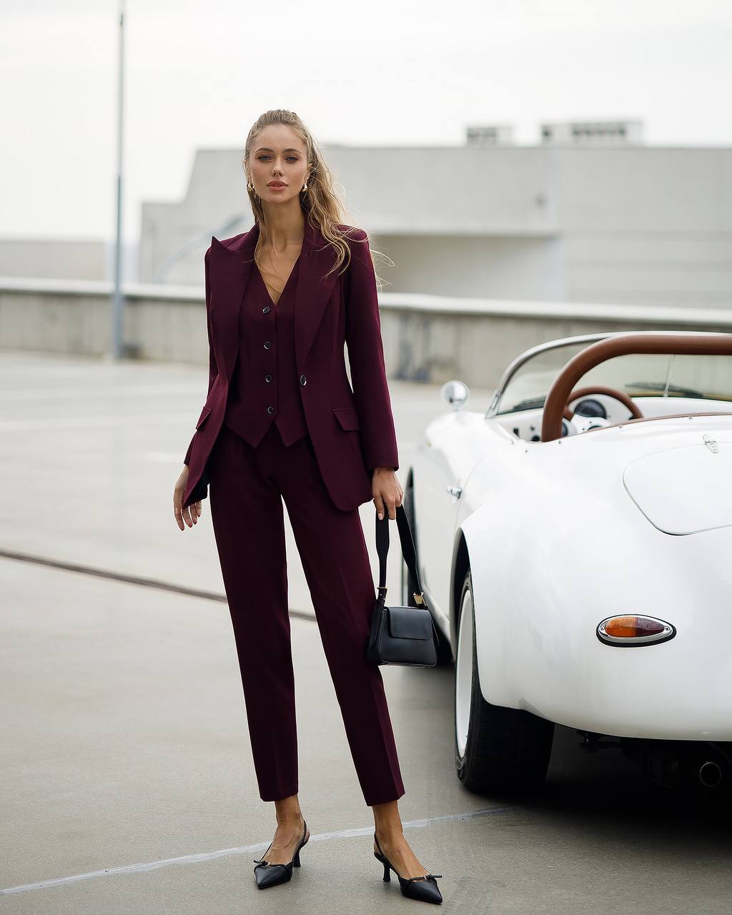 Woman in a burgundy suit standing next to a white vintage car on a road.