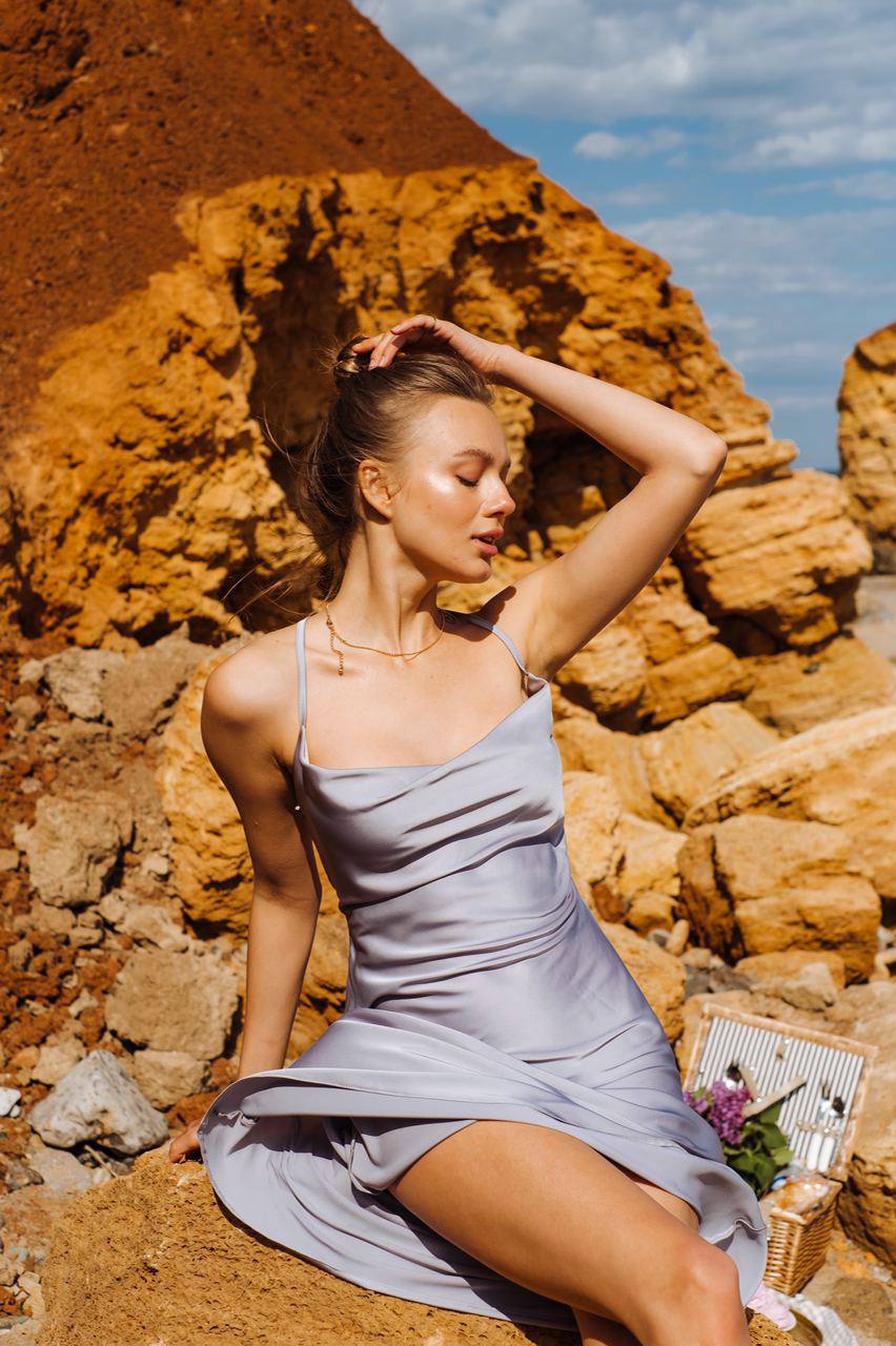 Woman in a light blue dress sitting on a rocky beach with a basket of flowers nearby.