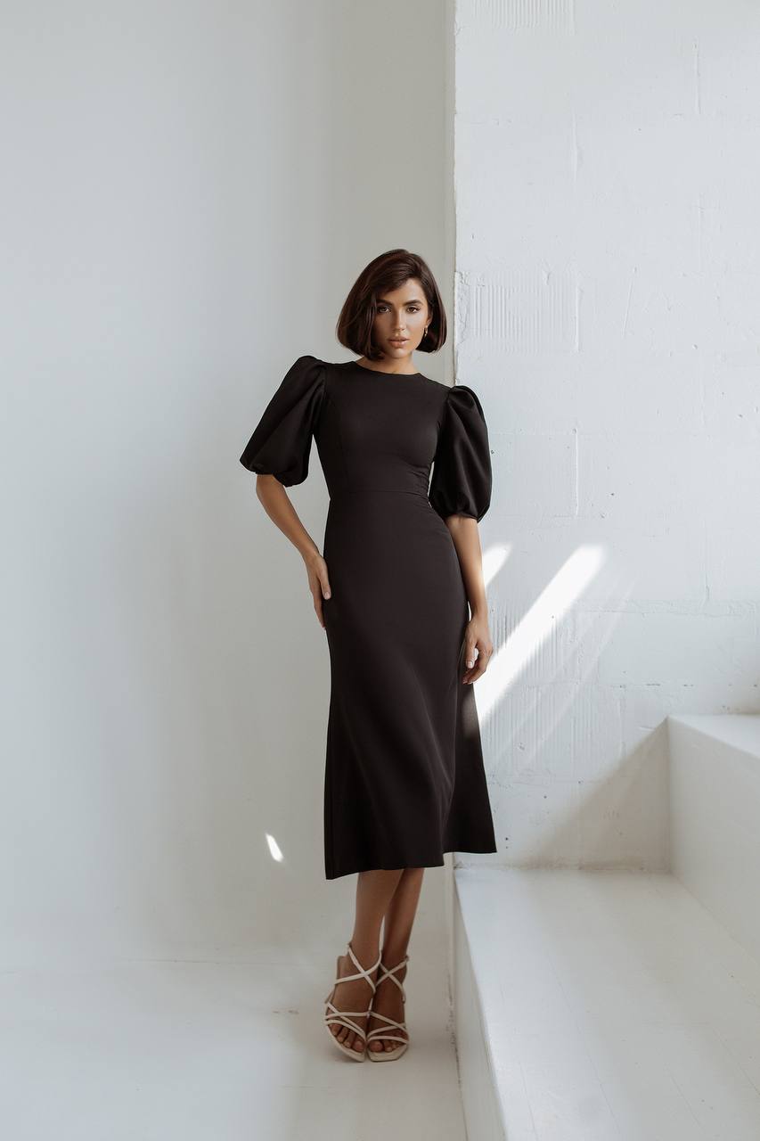 Woman wearing a black dress standing in a minimalistic room with white walls and floor.