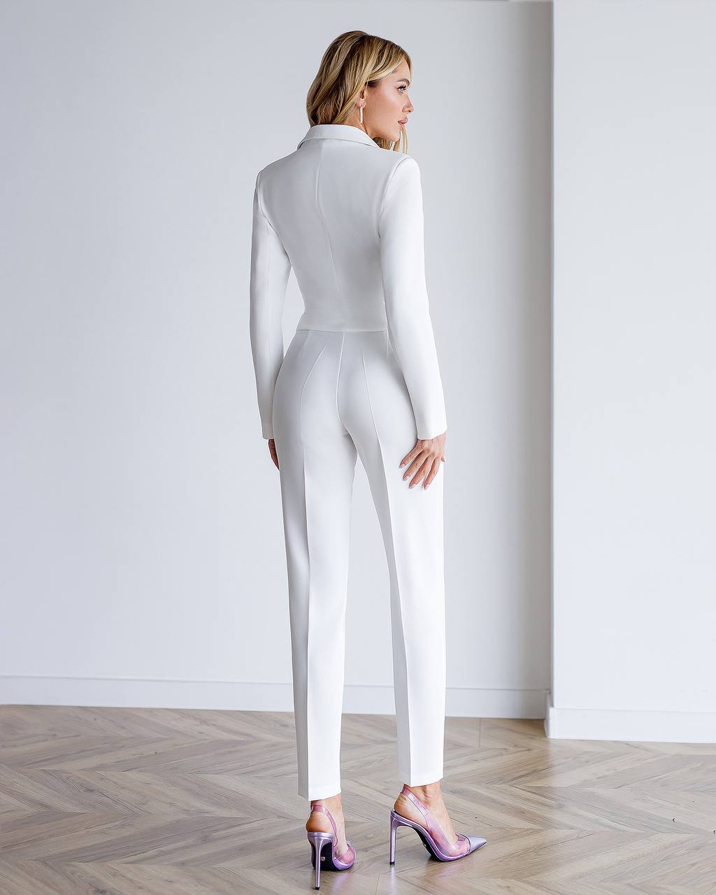 Woman wearing a white jumpsuit standing against a plain white wall.