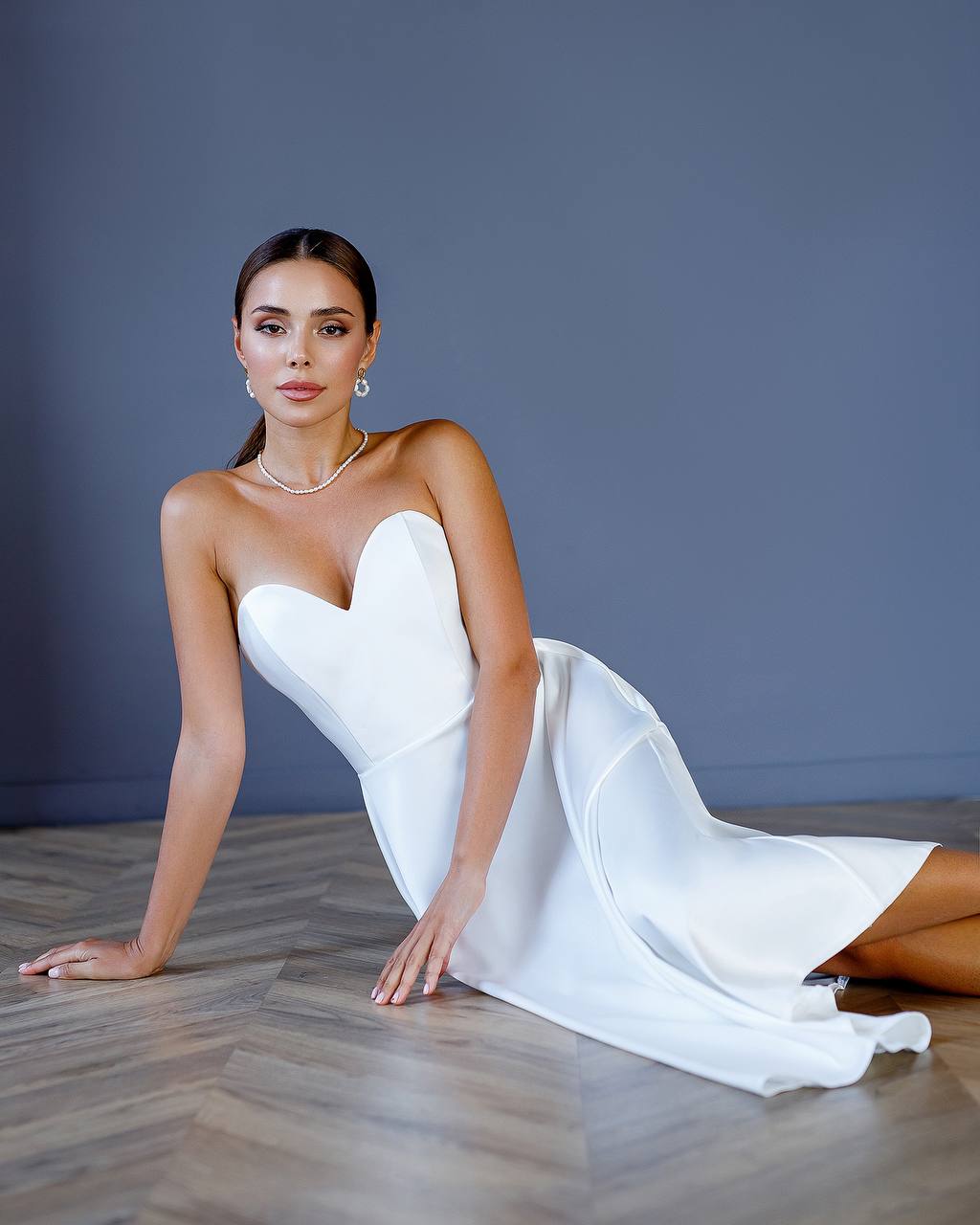 Woman in a white strapless dress sitting on a wooden floor against a gray wall.