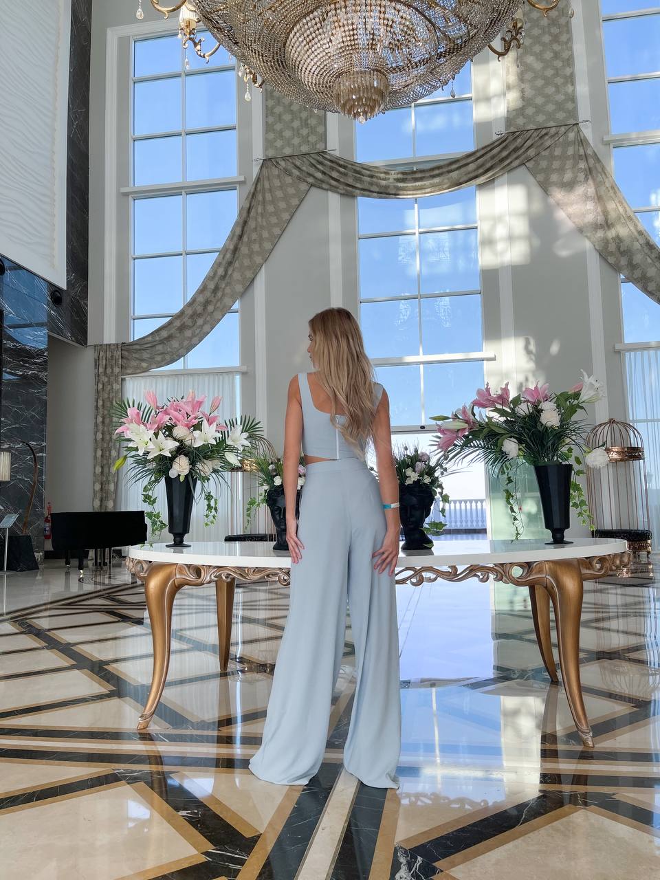 Woman in a light blue dress standing in a luxurious room with large windows and decorative flowers.