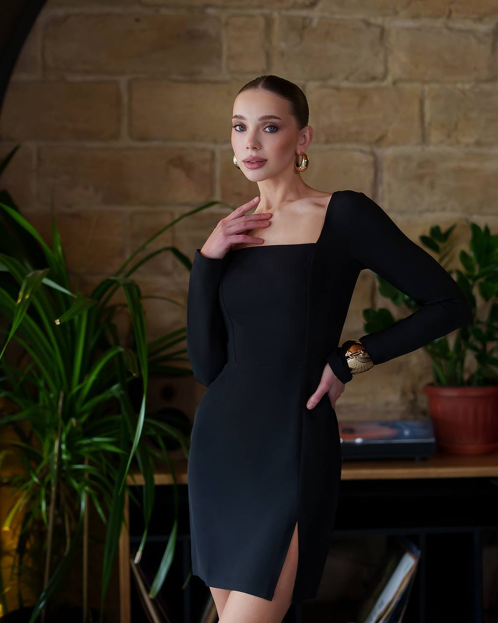 Woman in a black dress posing against a brick wall with plants.