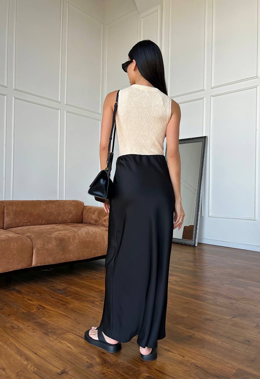 Woman in a beige top and black pants standing in a room with a brown sofa and white paneled walls.