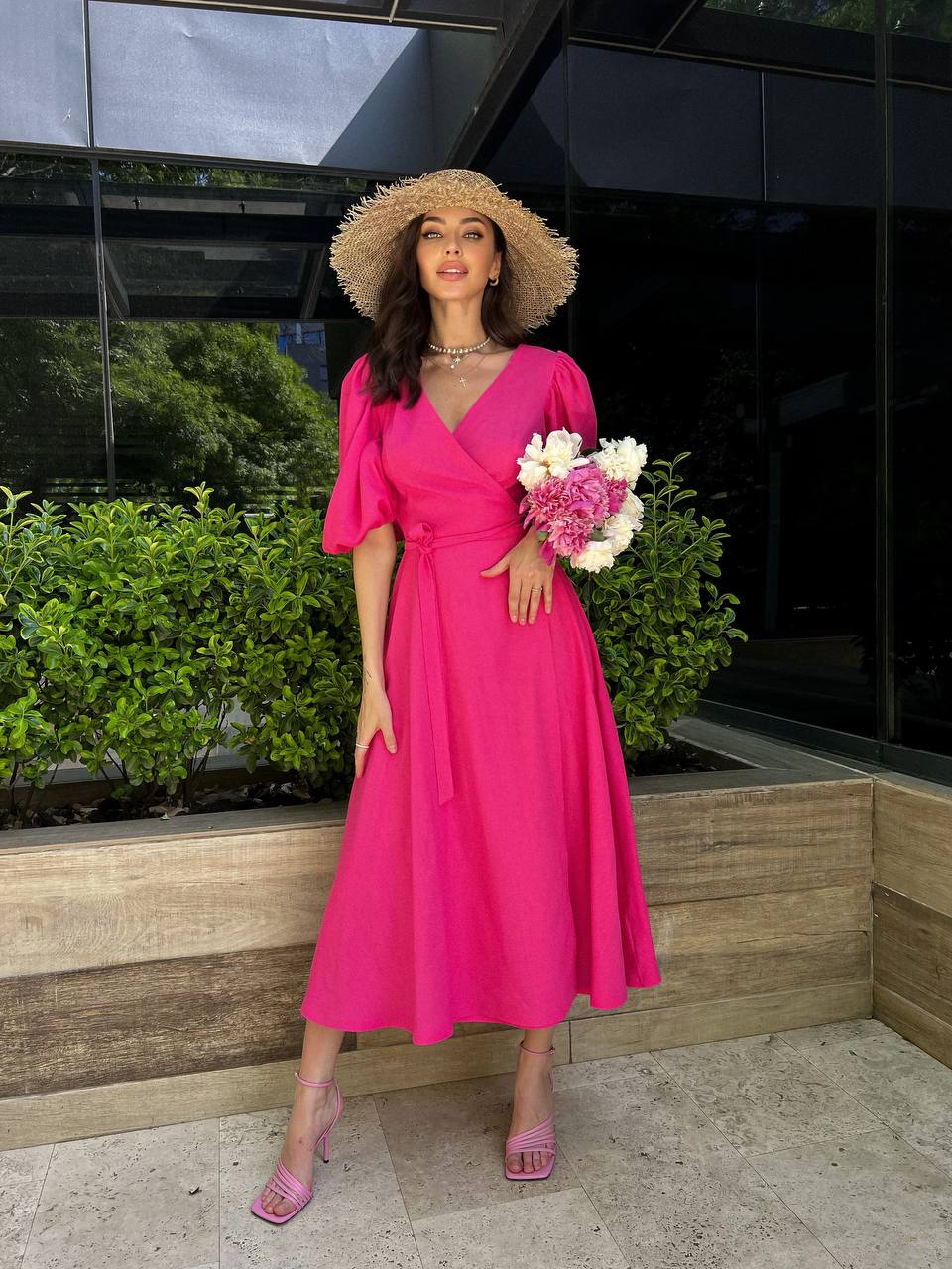 Woman in a bright pink dress and straw hat holding flowers, standing outdoors.