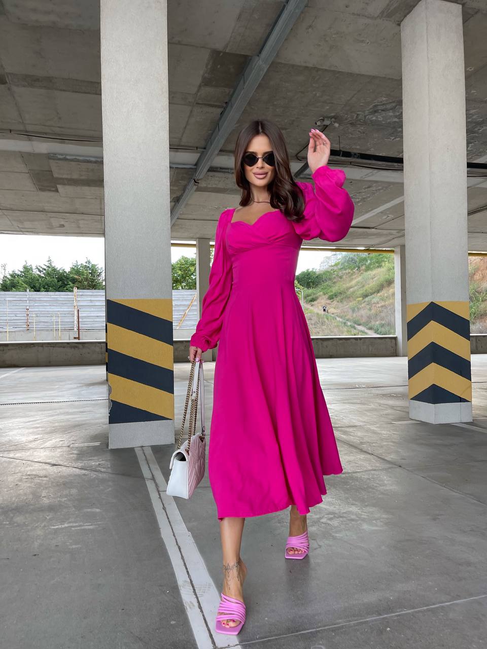 Woman in a bright pink dress standing under a bridge with concrete pillars and a road in the background.