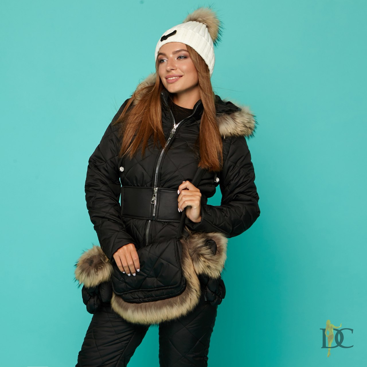 Woman wearing a black winter coat with fur trim and a white beanie against a teal background