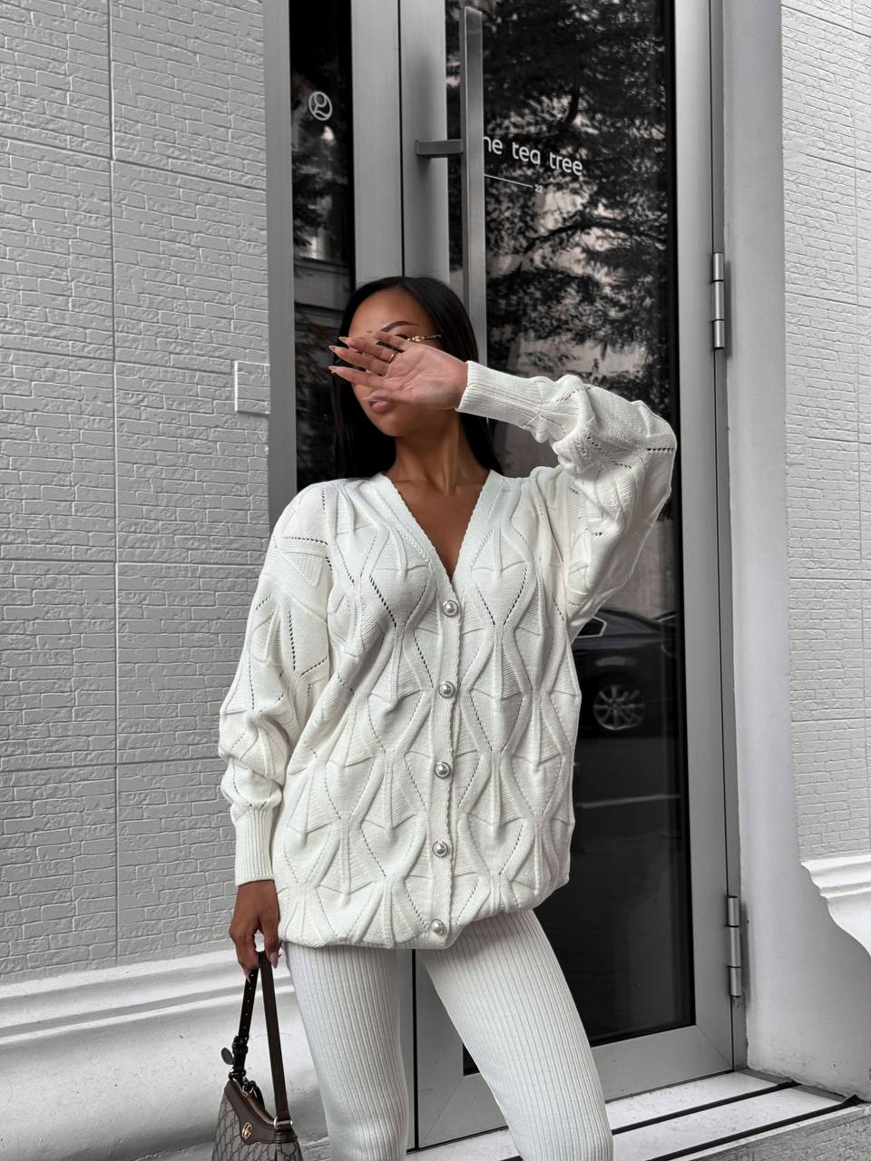 Woman wearing a white textured cardigan and pants standing in front of a glass door.