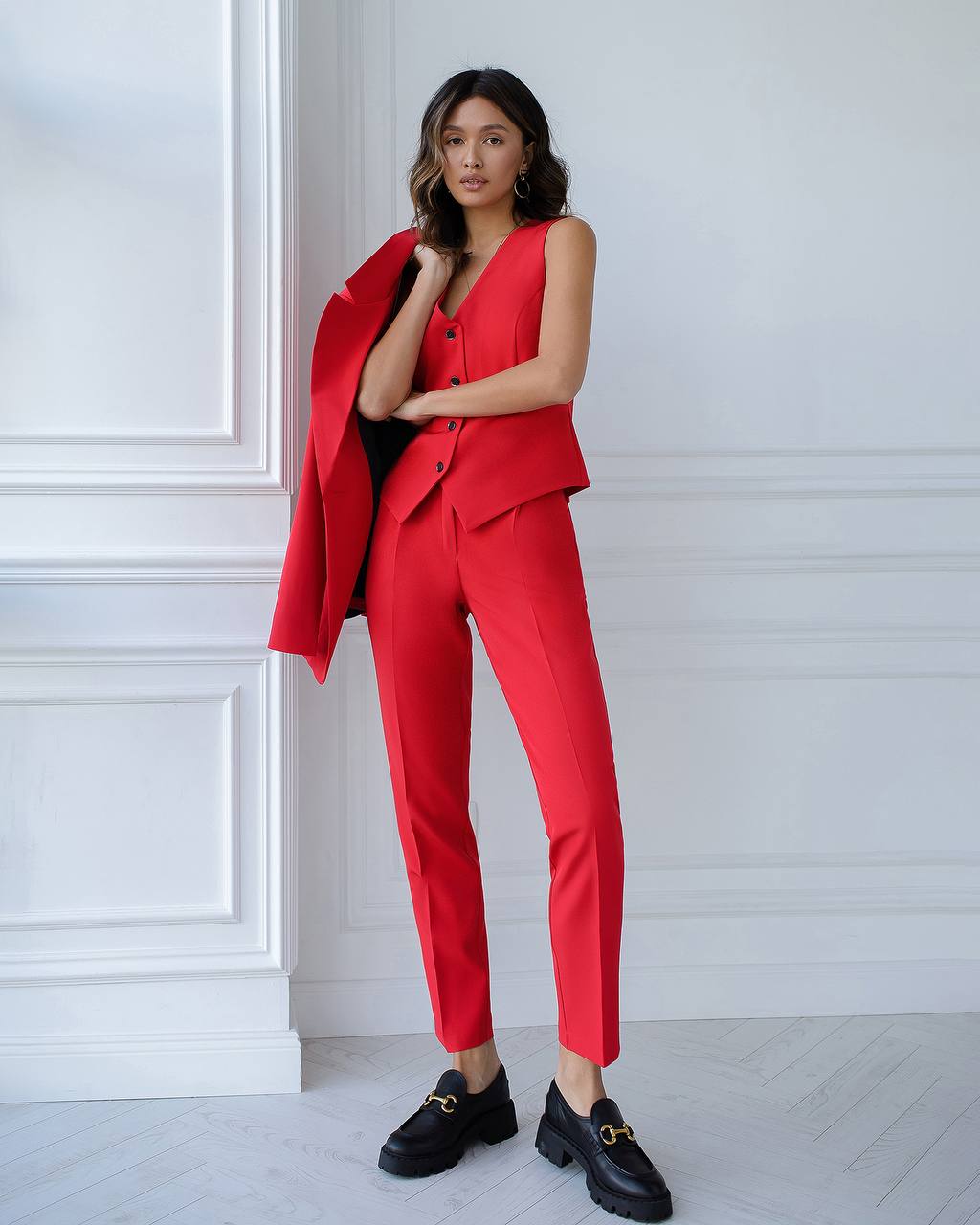 Woman wearing a red suit with black shoes against a white paneled wall.