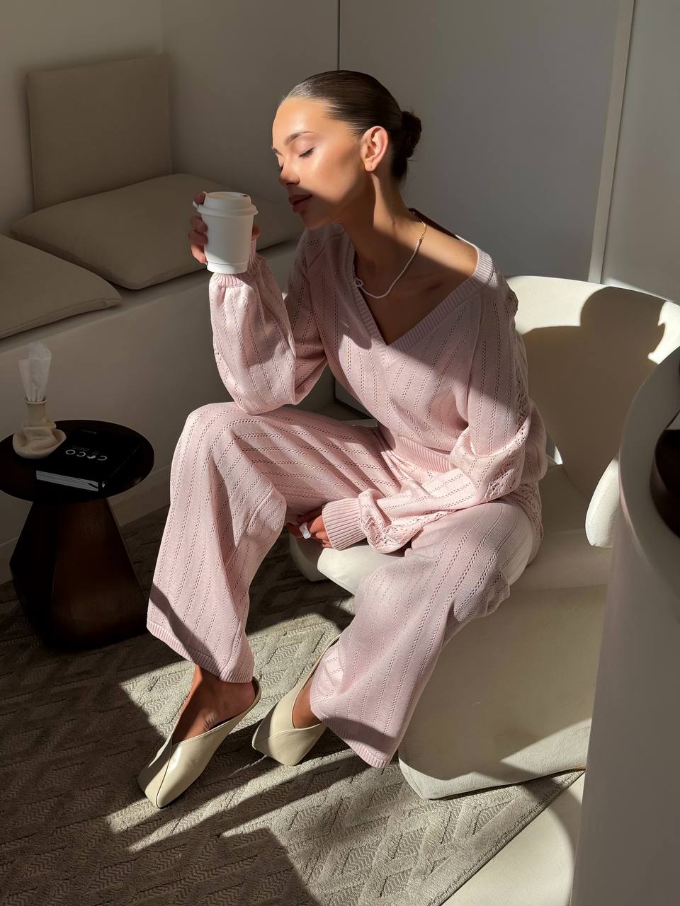 Woman in pink pajamas sitting on a chair holding a white cup in a cozy room.