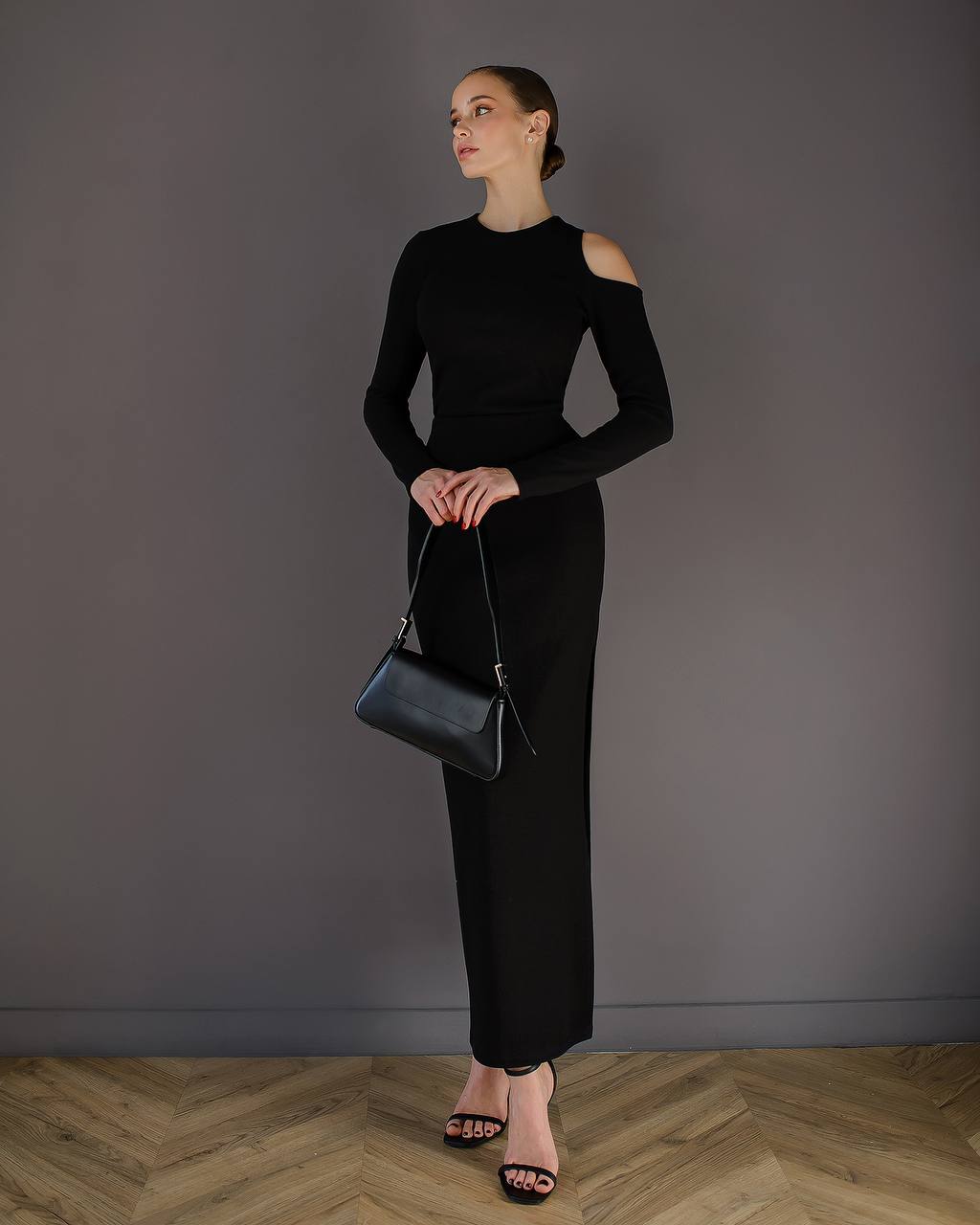 Woman in a black outfit holding a black handbag against a gray wall.