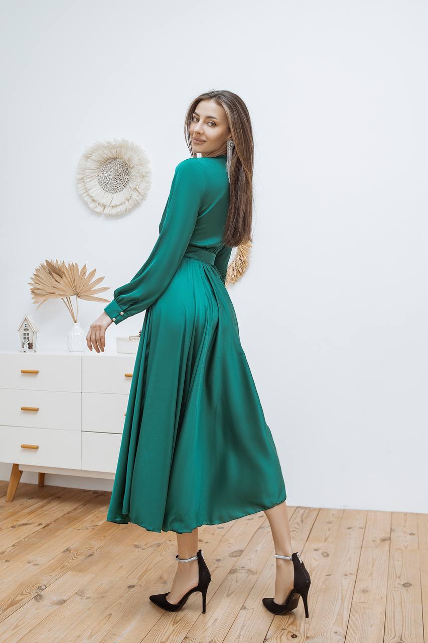Woman in a green dress standing in a room with a white wall and wooden floor.