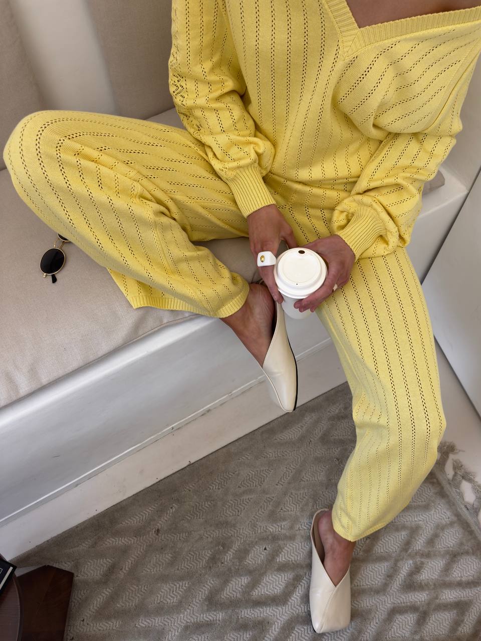 Person wearing a yellow knitted outfit sitting on a couch holding a white cup.