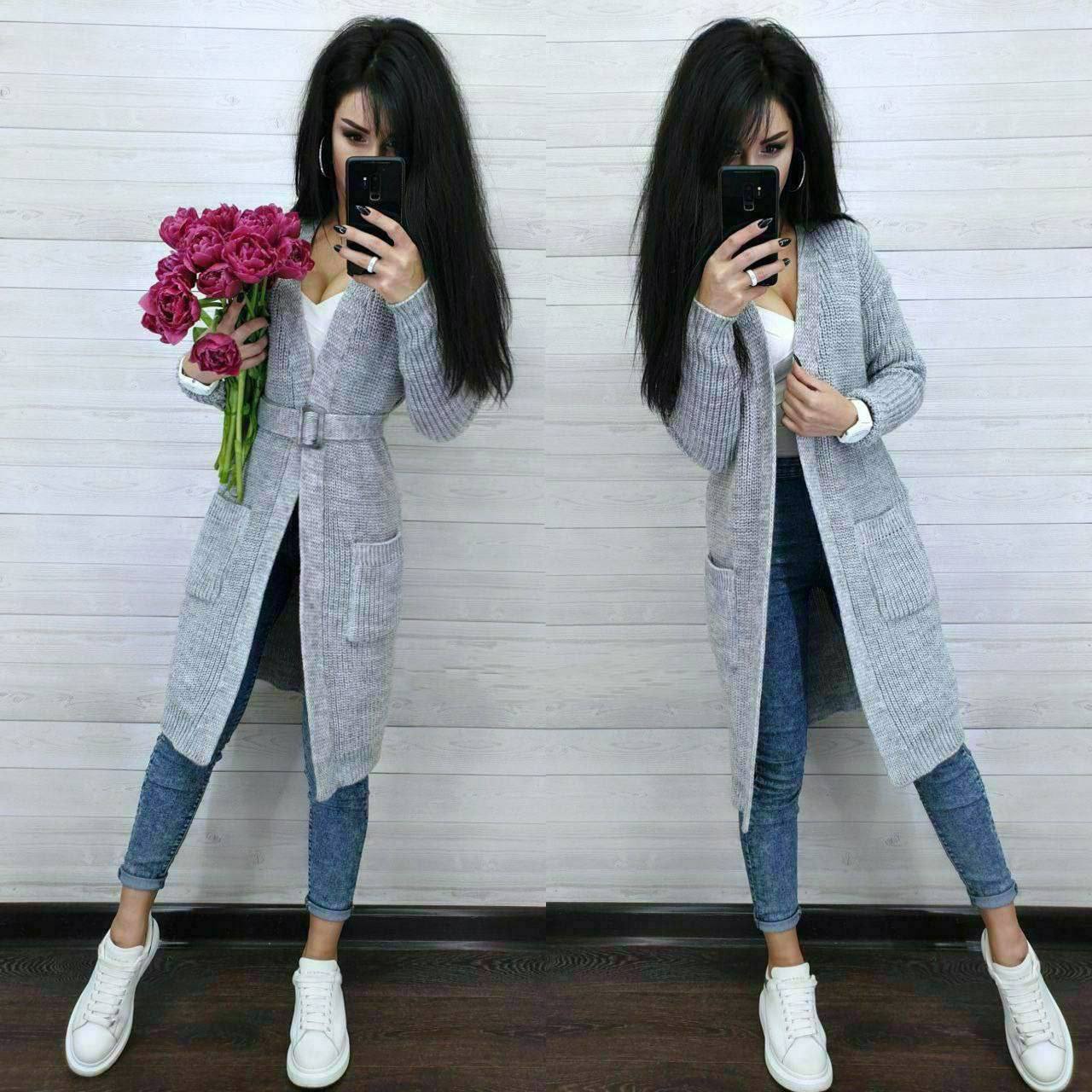 Woman wearing a long gray cardigan over a white shirt, blue jeans, and white sneakers, holding pink flowers.