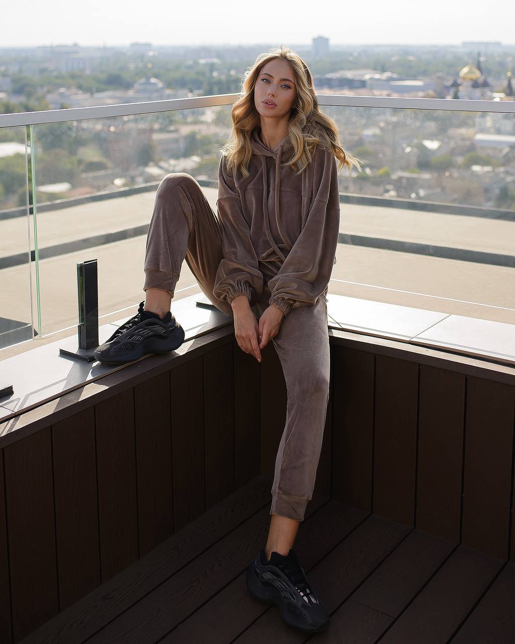 Woman in a brown sweatshirt and pants sitting on a rooftop with a cityscape view.