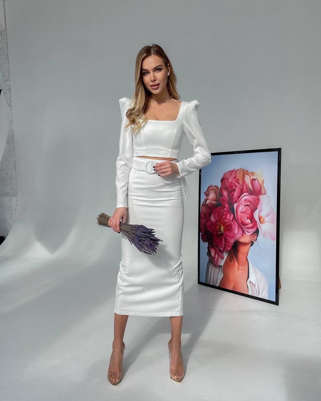 Woman in a white outfit holding flowers, standing next to a framed artwork on a gray background