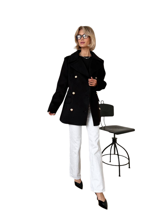 Woman wearing a black coat and white pants standing next to a black chair on a white background