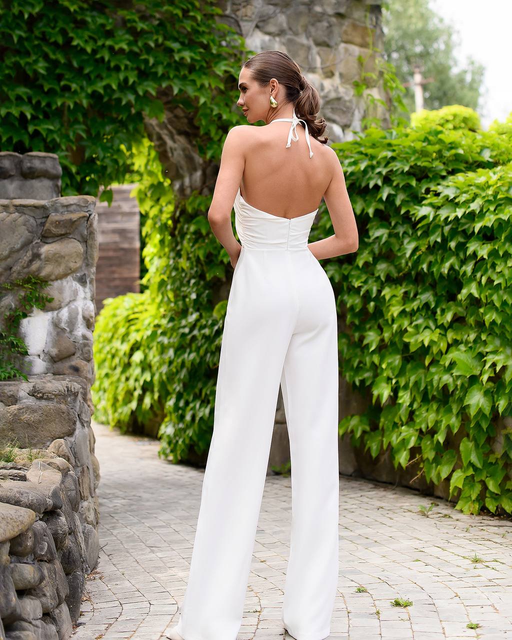 Woman in a white jumpsuit standing in a garden with greenery and stone walls.