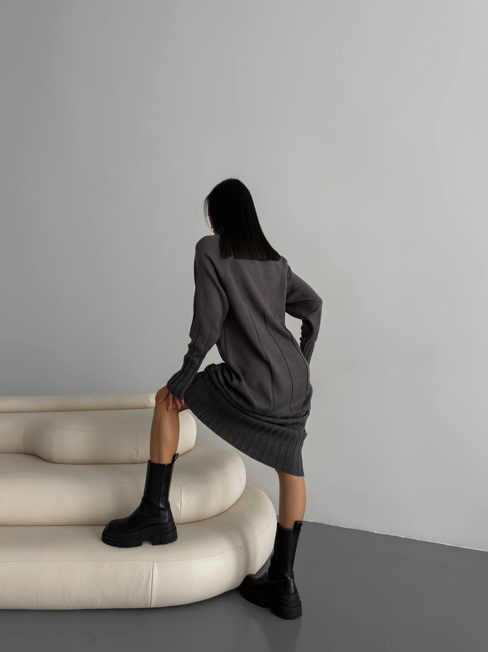 Woman in a gray dress sitting on a white curved sofa against a plain wall.