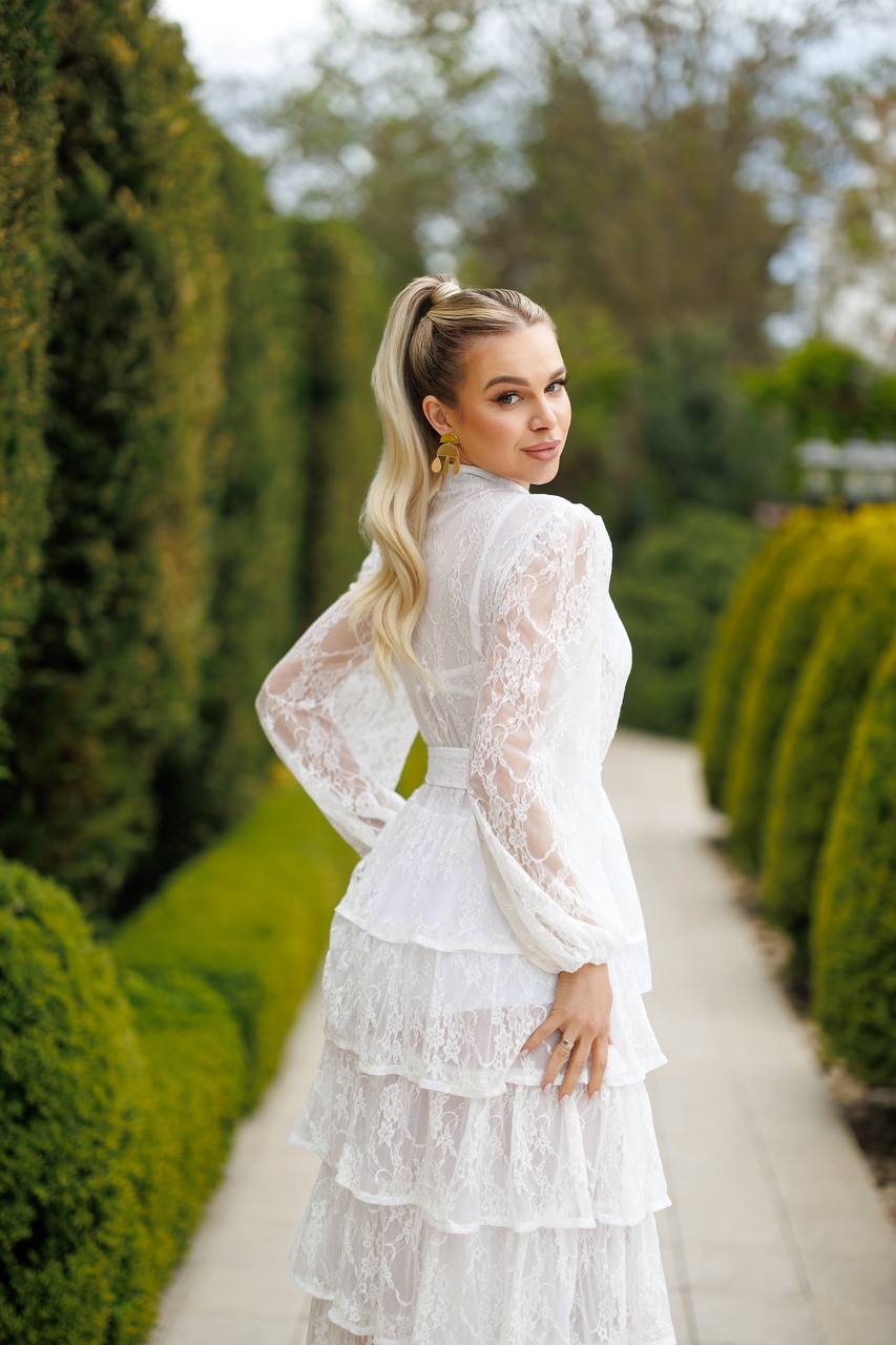 Woman in a white lace dress standing in a garden with green hedges.