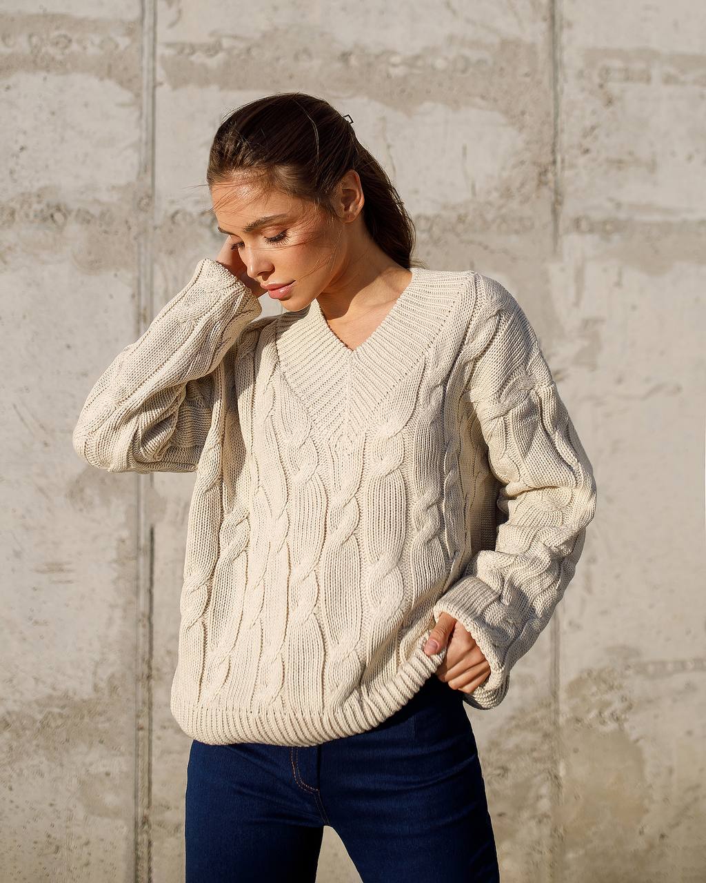 Woman wearing a cream cable knit sweater against a textured wall.