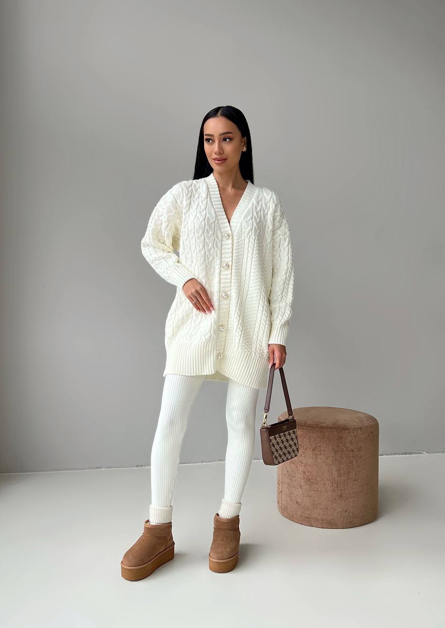 Woman wearing a white cardigan, white pants, and brown boots holding a small bag against a gray background.