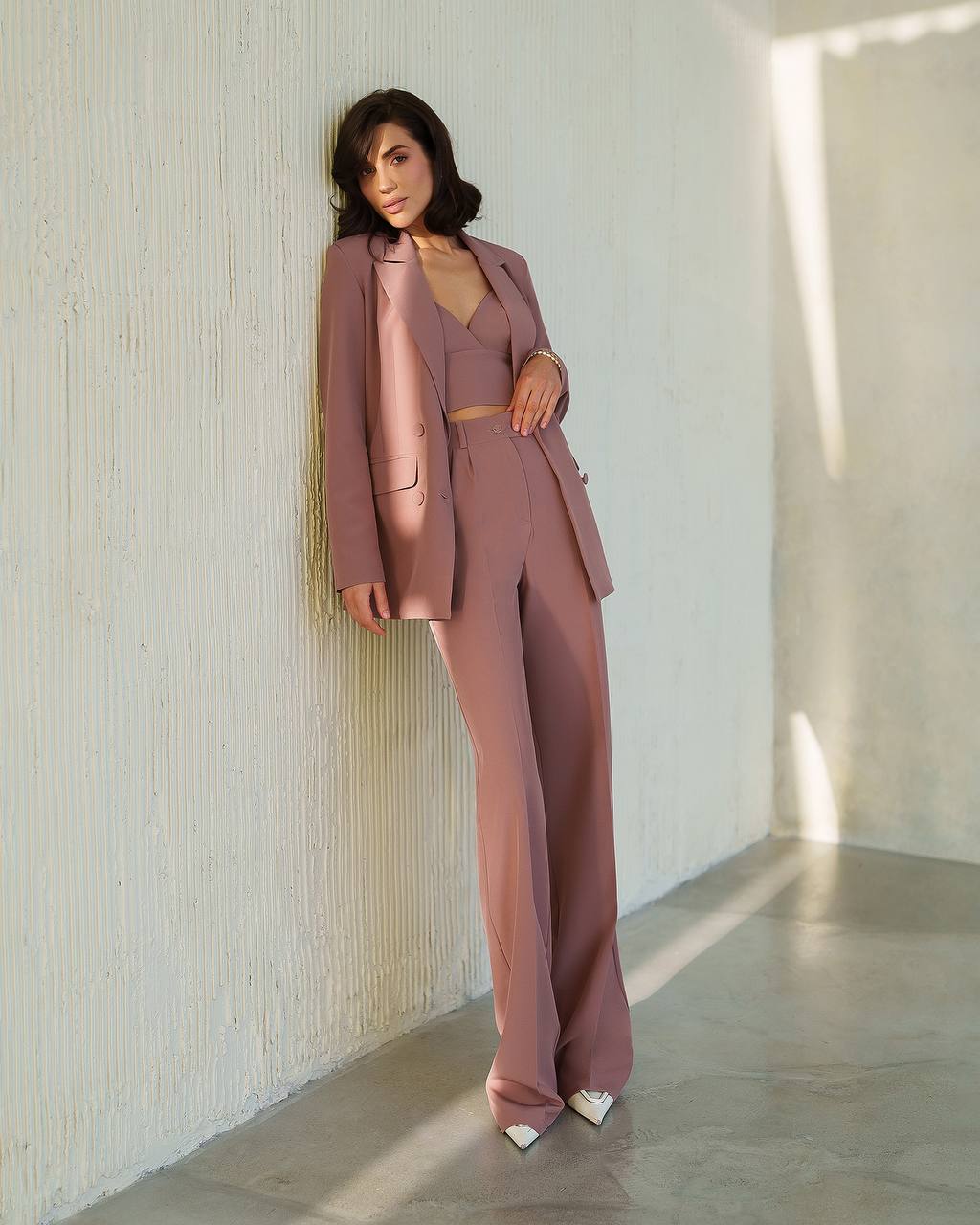 Woman in a pink suit standing against a textured white wall.