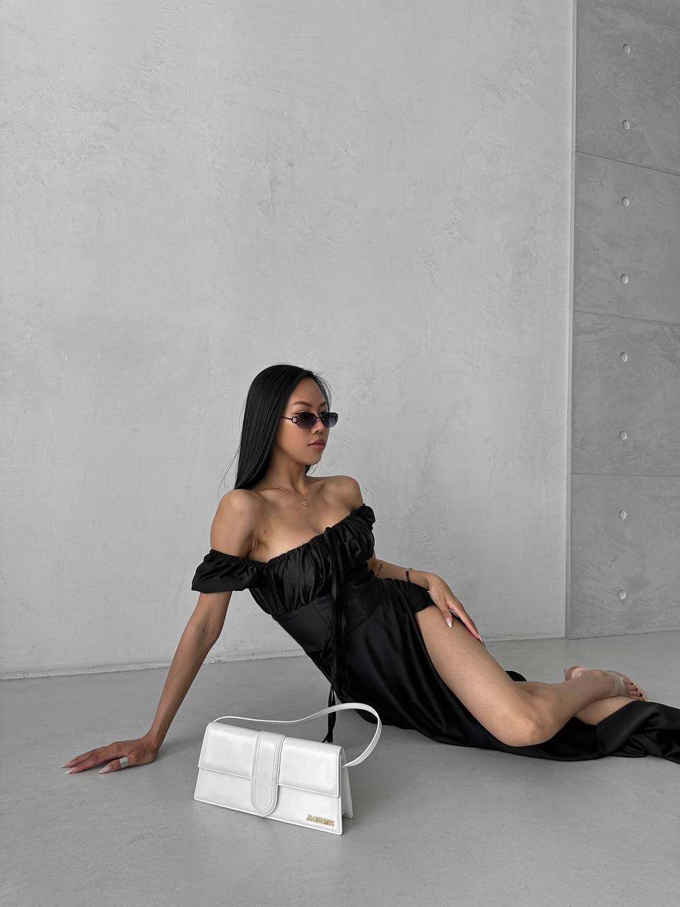 Woman in a black off-shoulder dress sitting on the floor with a white handbag against a gray concrete wall.