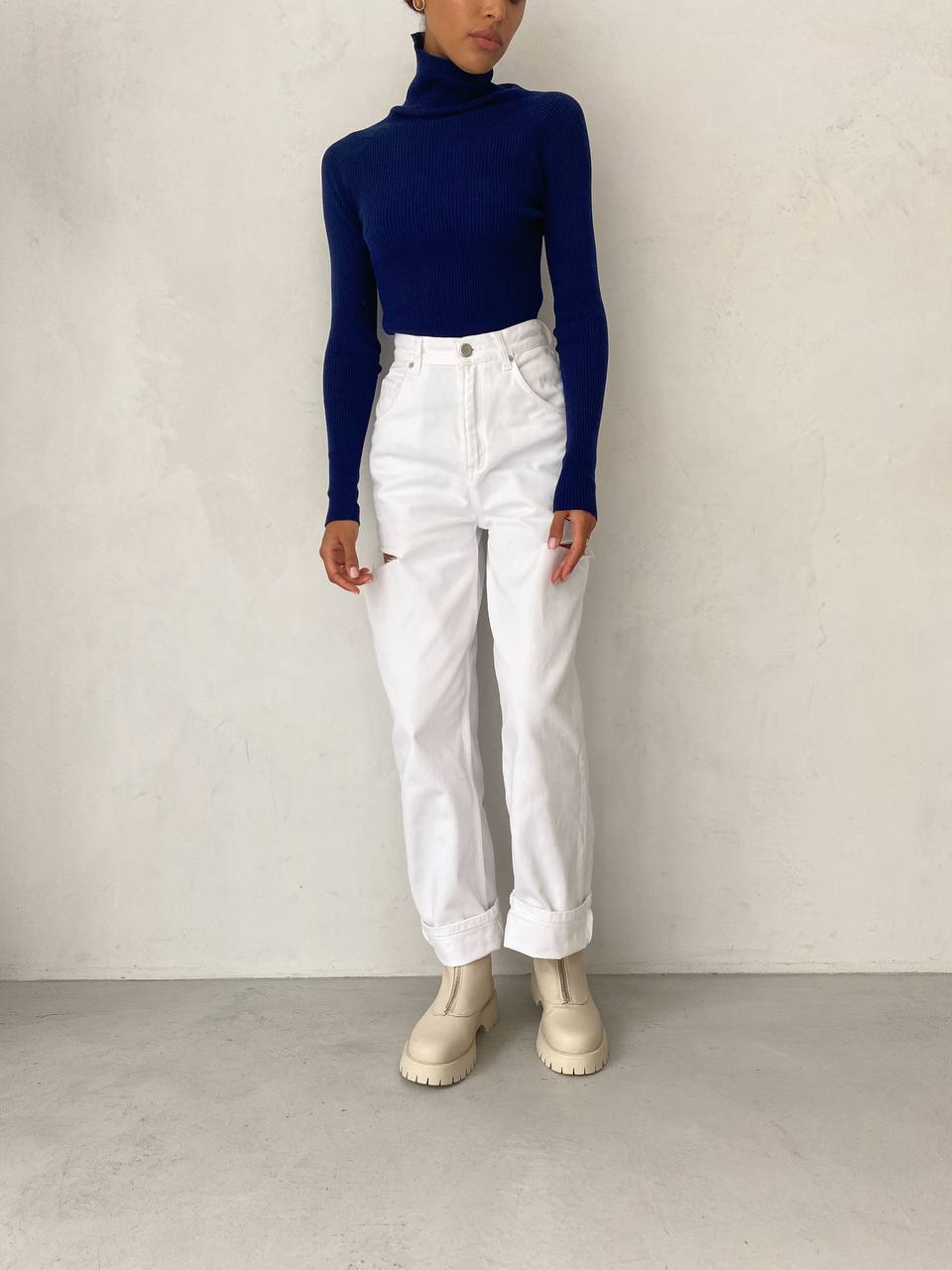 Person wearing a navy turtleneck, white jeans, and white boots against a plain background