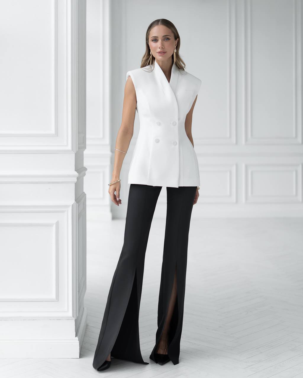 Woman wearing a white sleeveless top with cut-out design and black pants against a white paneled wall.