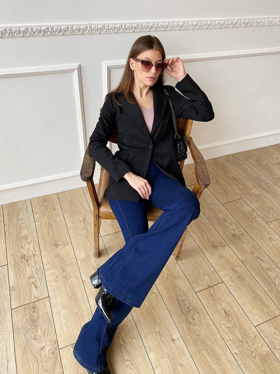 Woman sitting on a chair wearing a black blazer and blue jeans in an elegant room.