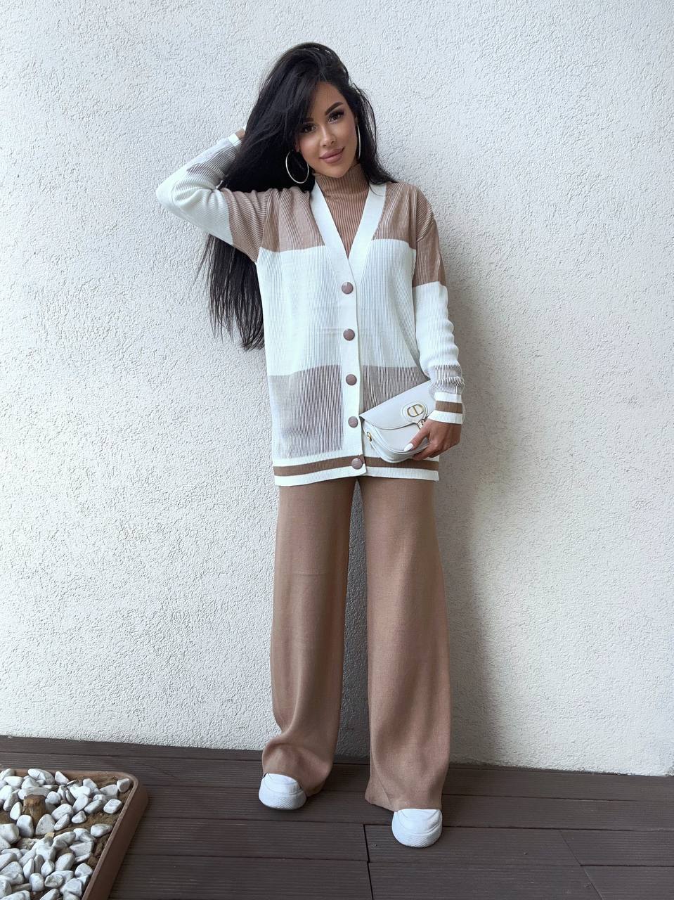 Woman wearing a white and beige cardigan with matching pants against a plain wall.