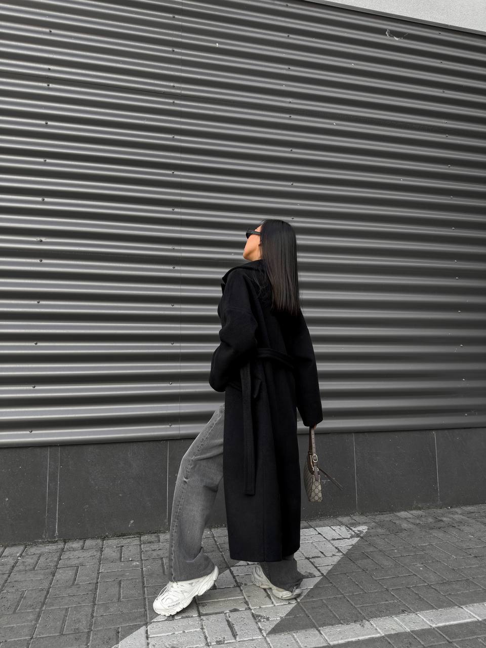 Person wearing a long black coat and light-colored pants standing against a gray corrugated metal wall.