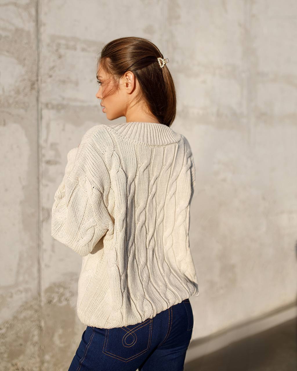 Woman wearing a cream cable knit sweater against a textured wall.