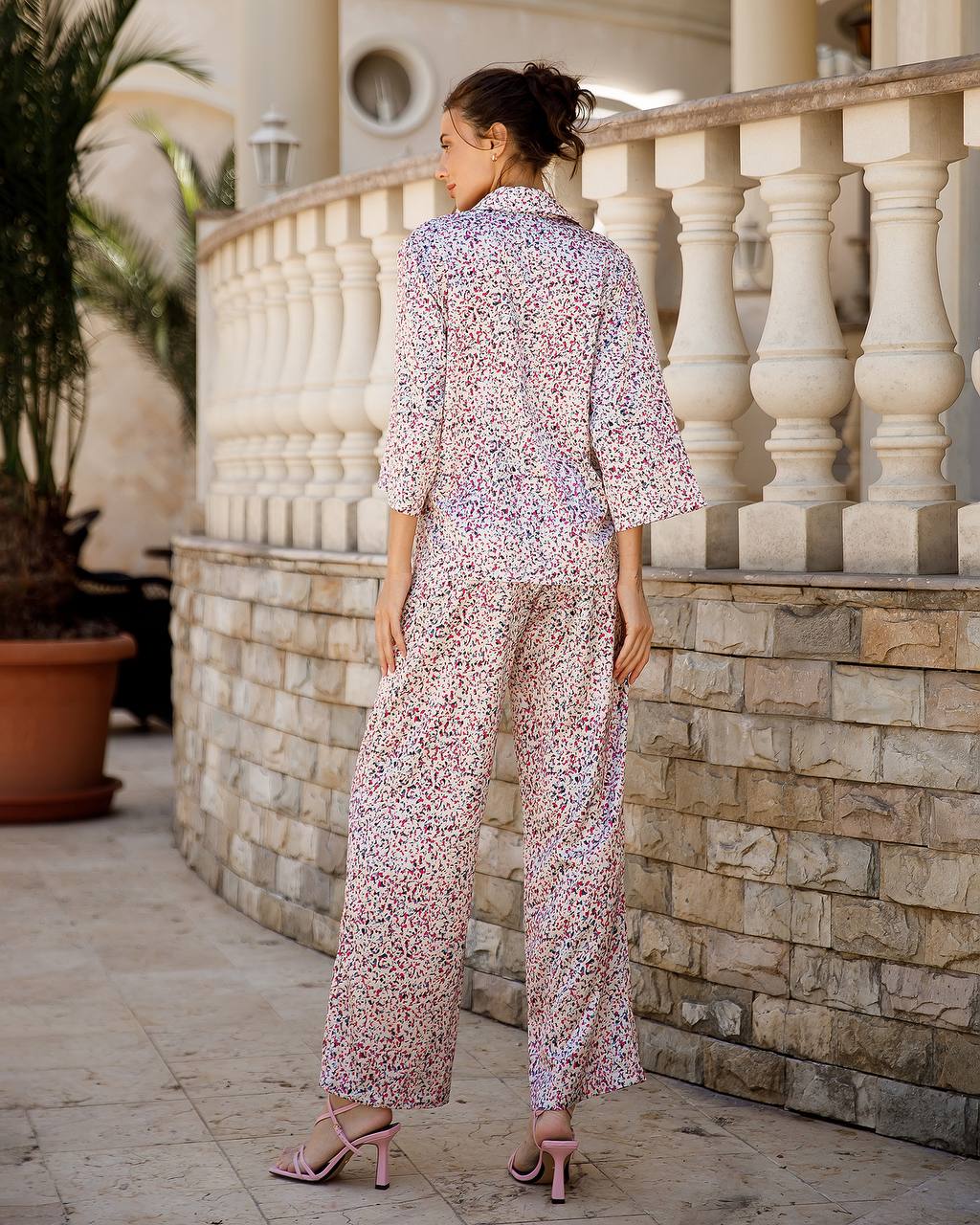 Woman in a floral outfit standing on a stone patio with a stone wall and railing in the background.