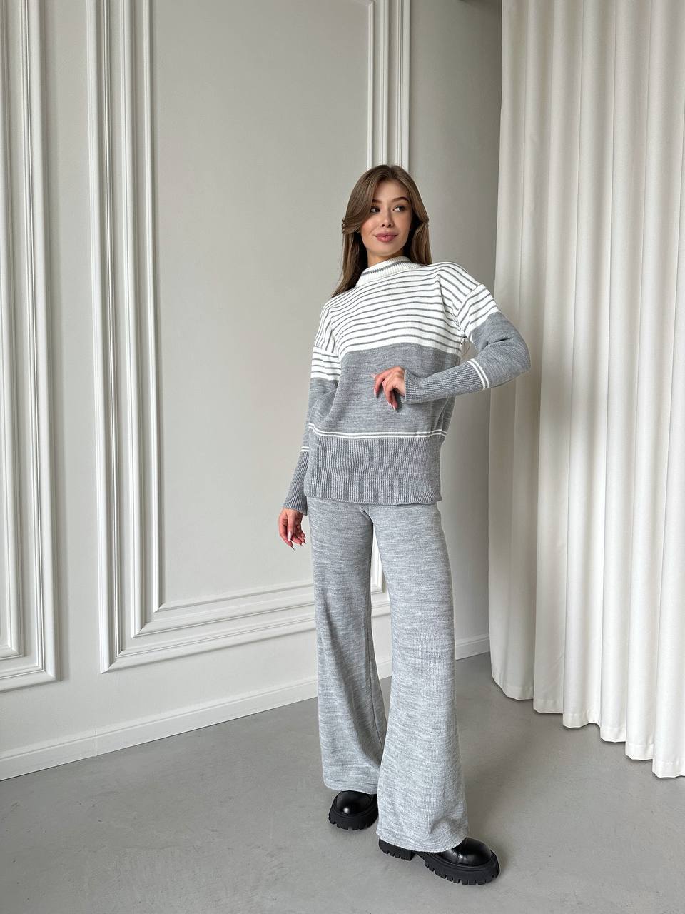 Woman wearing a gray and white striped sweater and pants in a room with white walls and curtains.
