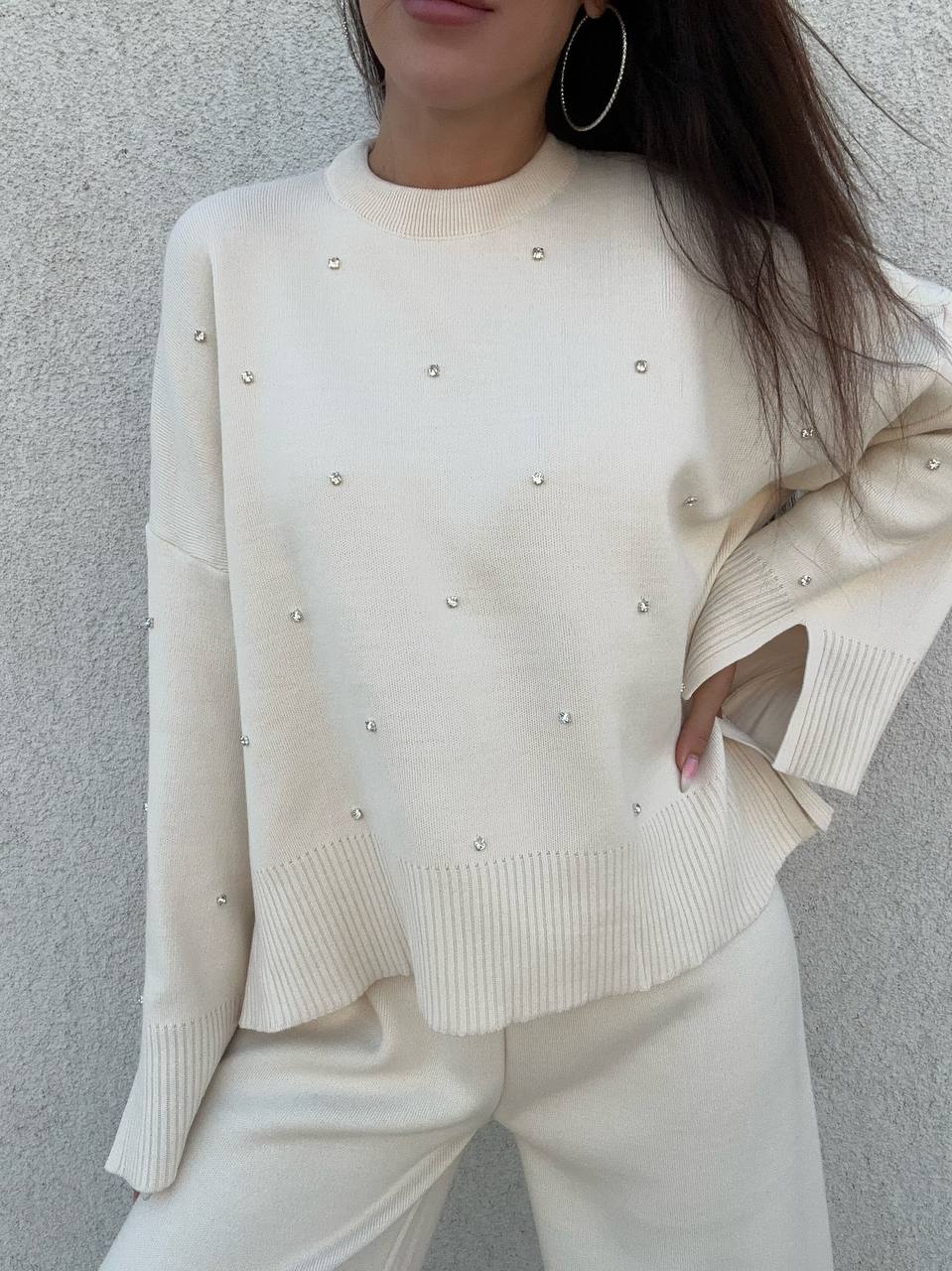 Person wearing a cream-colored sweater with pearl embellishments against a gray background