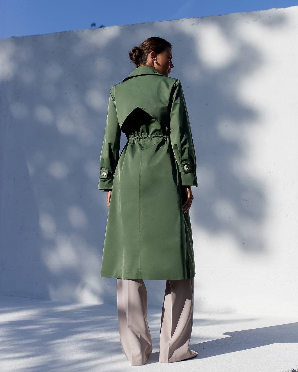 Person wearing a green trench coat standing against a white wall with blue sky.