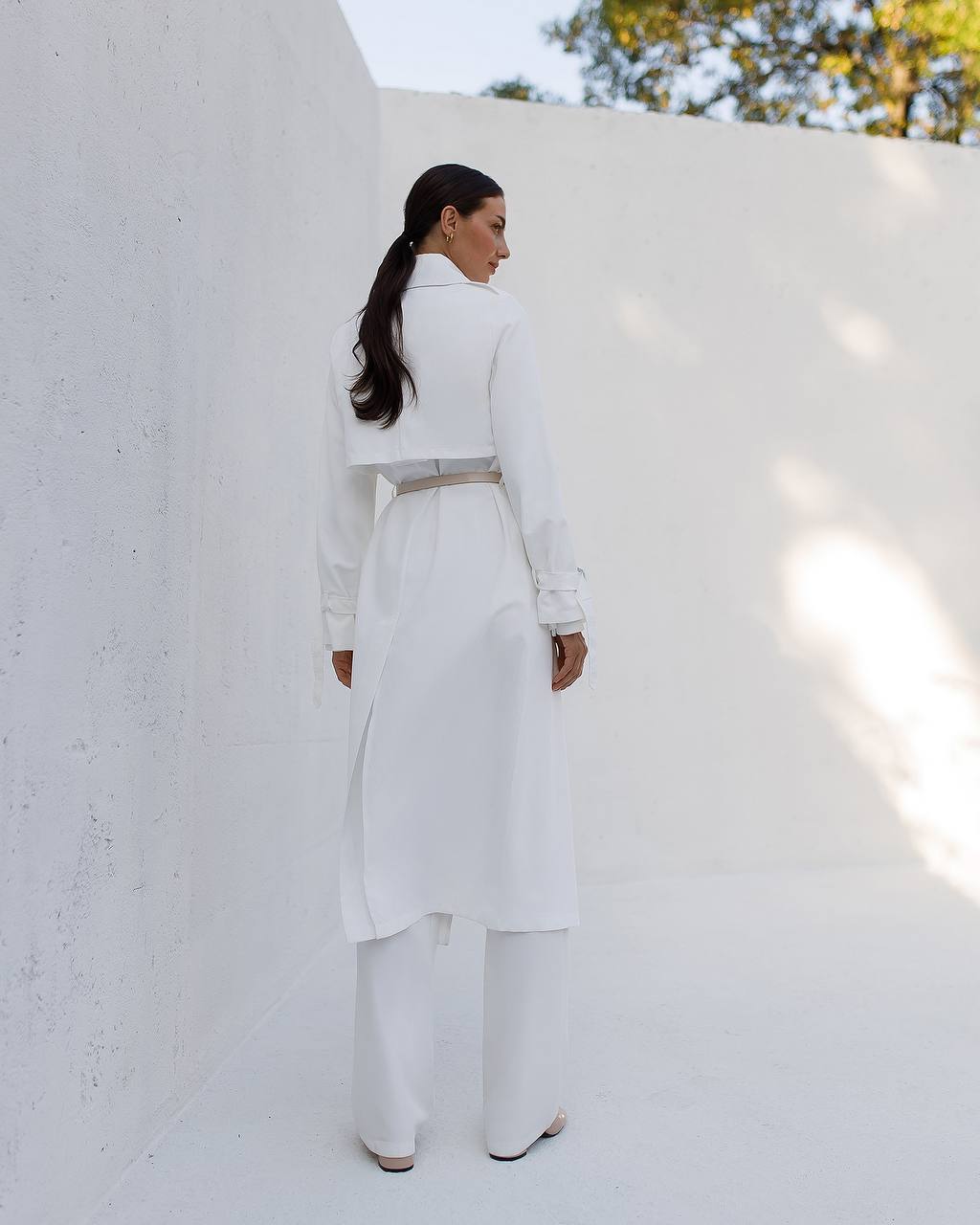 Person wearing a white outfit standing against a light-colored wall with trees in the background
