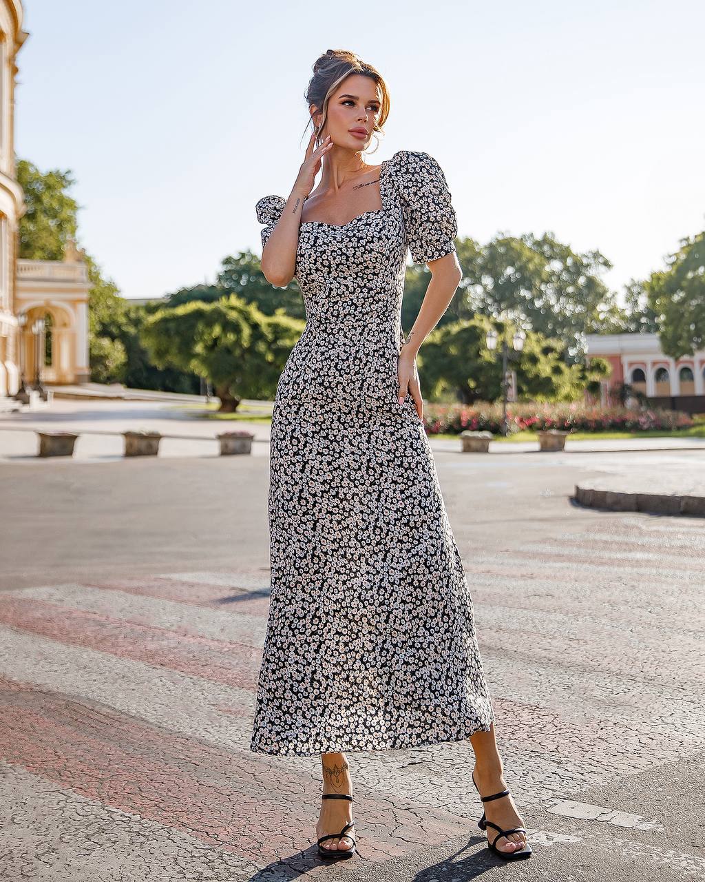 Woman in a floral dress standing outdoors with trees and buildings in the background