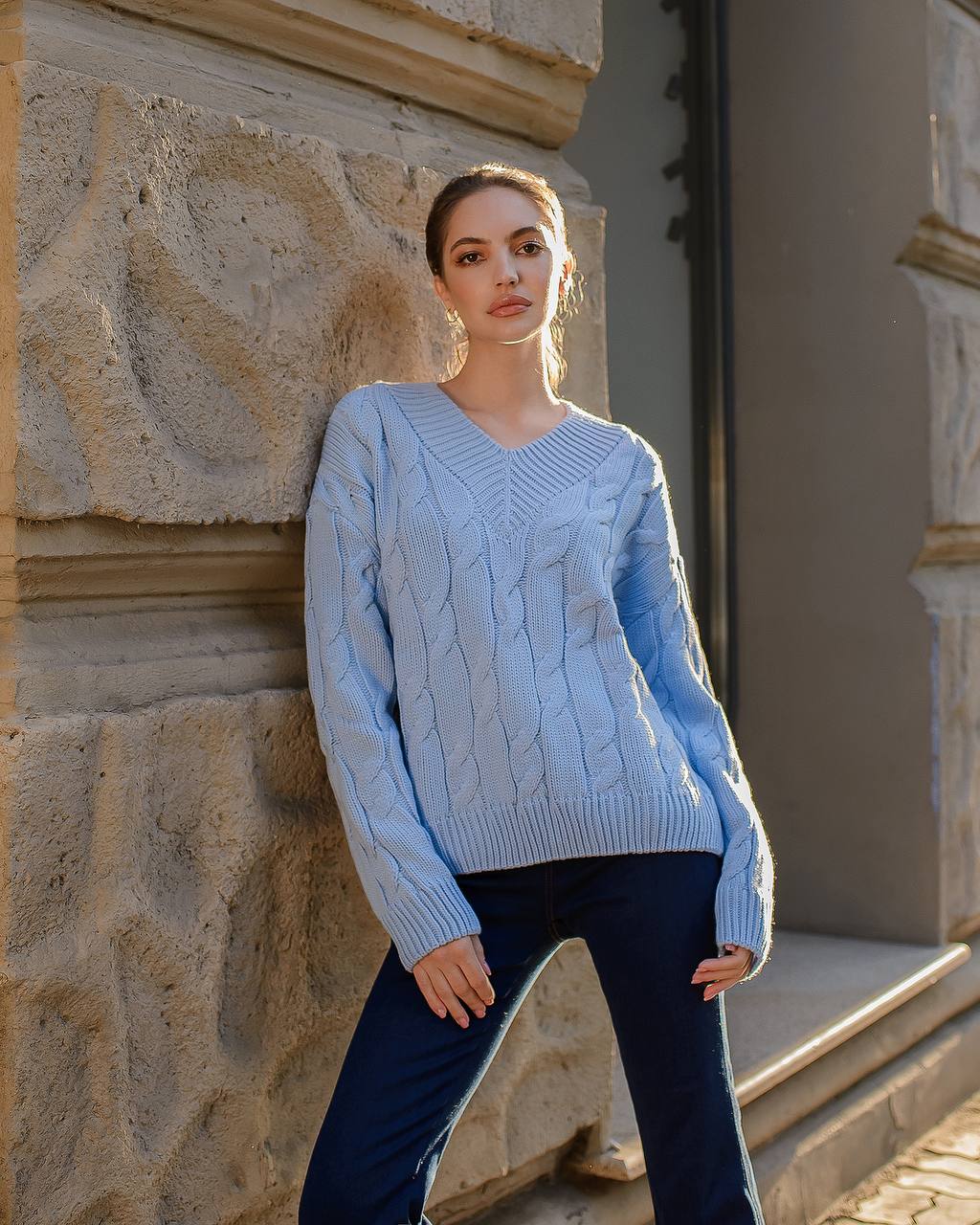 Woman wearing a light blue cable knit sweater standing against a stone wall.