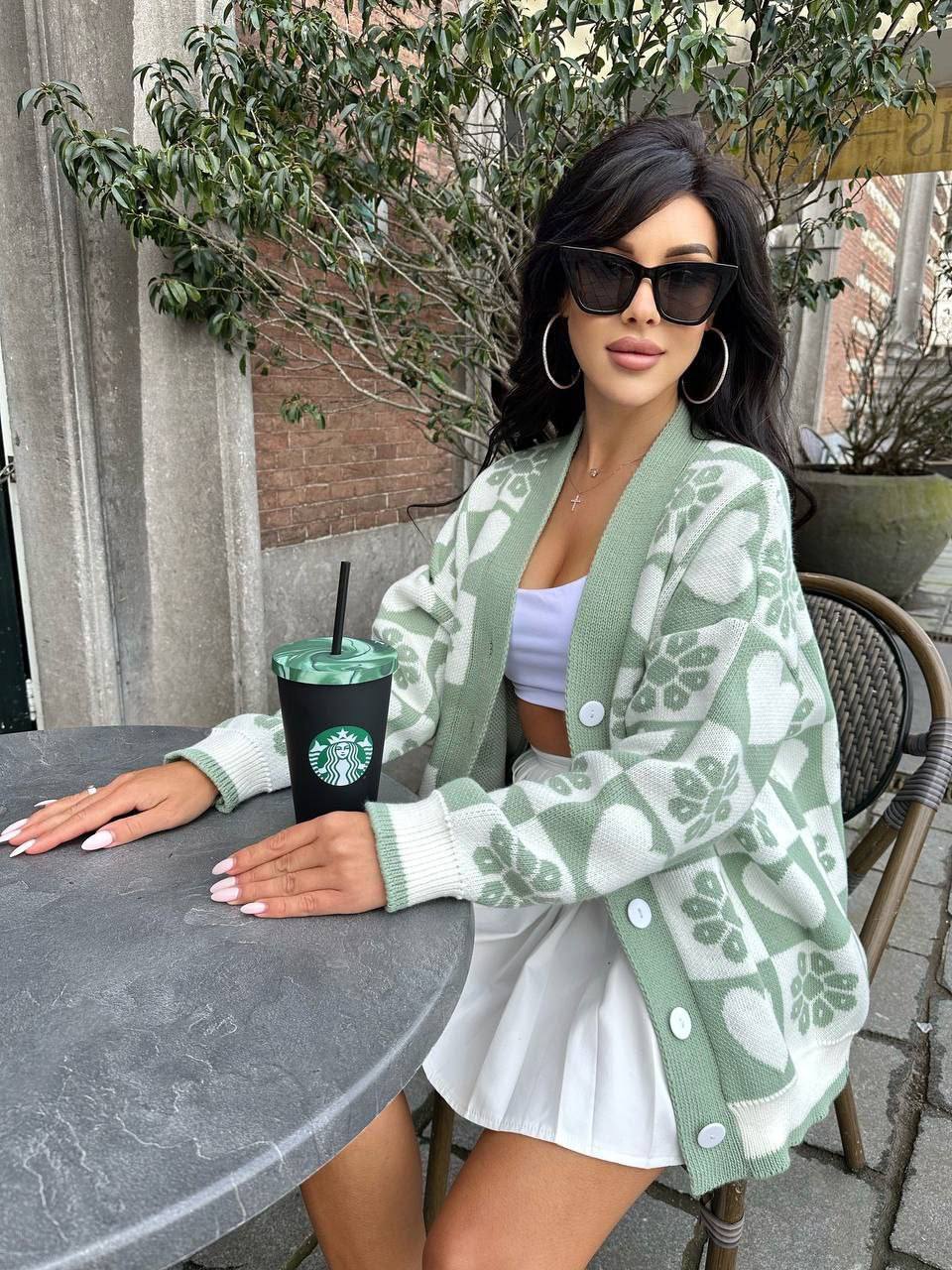 Woman in a green cardigan with white patterns sitting outdoors, holding a Starbucks cup.
