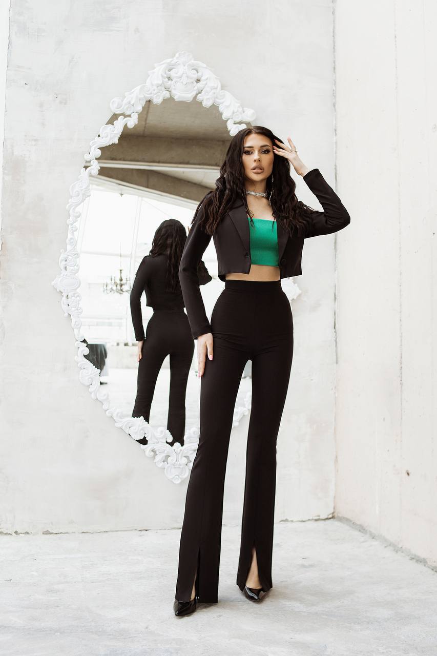 Woman in black outfit with green top standing in front of a decorative mirror.