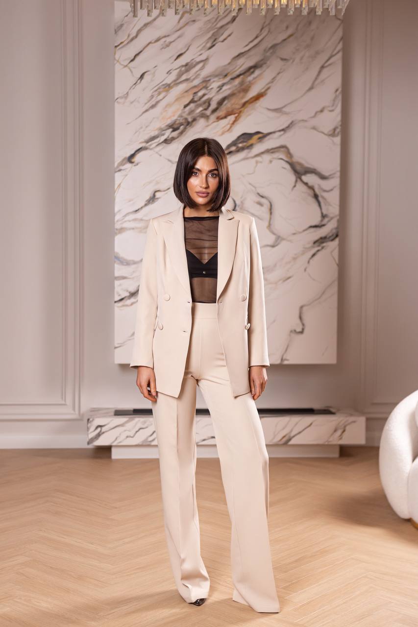 Woman in a beige suit standing in a room with marble wall and wooden floor.
