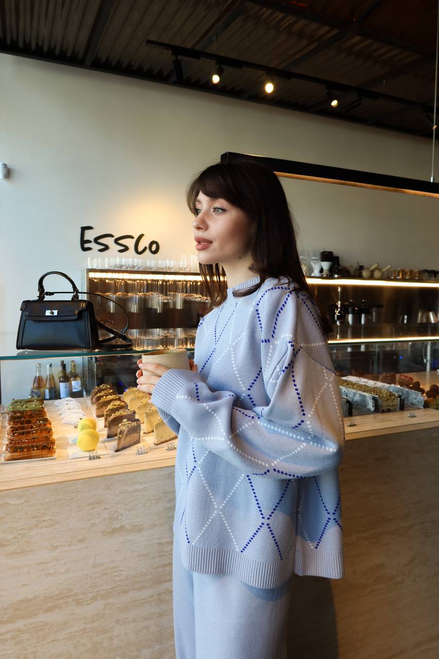 Woman in a cafe wearing a light blue sweater with a patterned design, standing in front of a display case with various items.