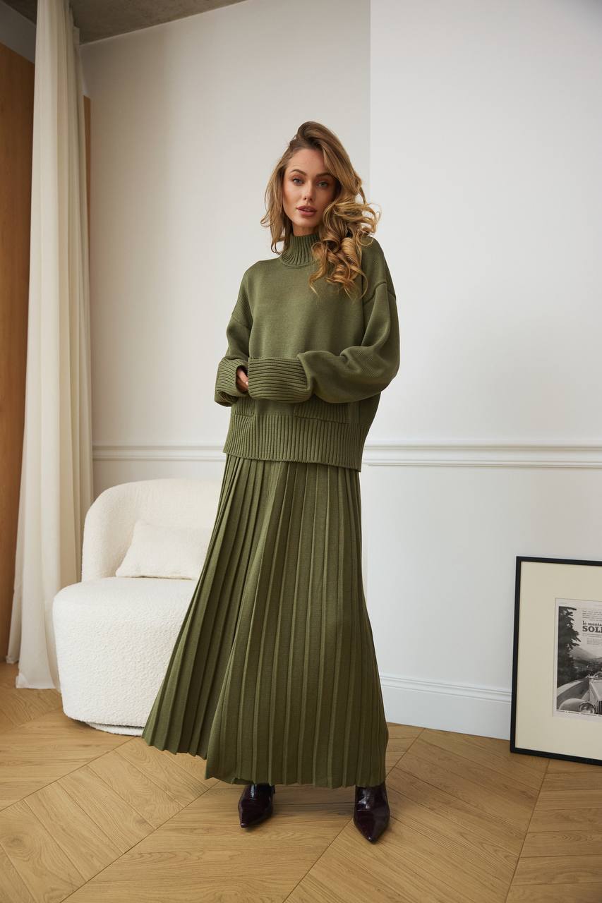 Woman wearing a green sweater and pleated skirt standing in a room with a white wall and a framed picture.