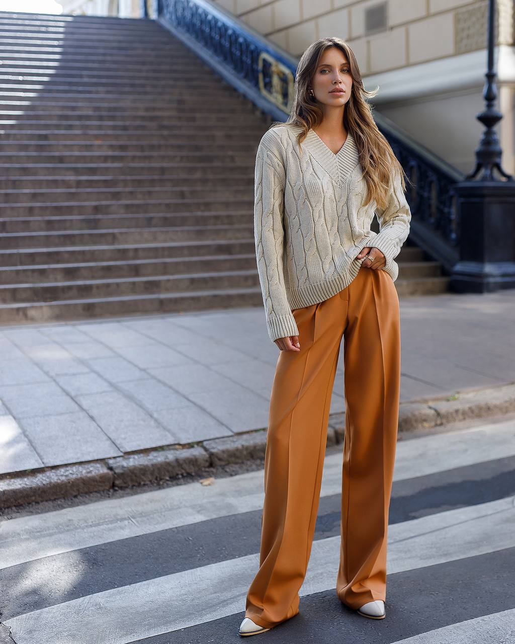 Woman in a cream sweater and brown pants standing on a city street.